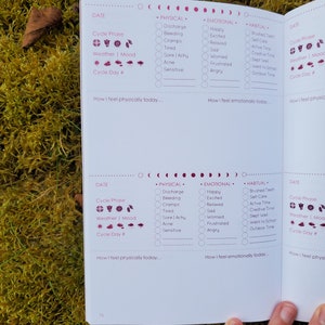 May include: A two-page spread from a pink and white journal with a section for tracking physical, emotional, and habitual activities. The journal includes sections for tracking cycle phase, weather, and mood. The journal has a pink and white color scheme with a pink moon and sun graphic.