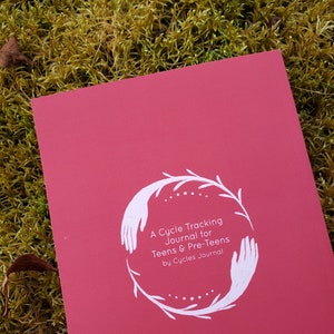 May include: A red journal with a white illustration of hands holding a wreath of leaves. The text on the cover reads "A Cycle Tracking Journal for Teens & Pre-Teens by Cycles Journal."