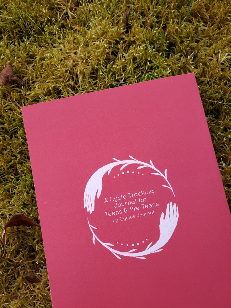 May include: A red journal with a white illustration of hands holding a wreath of leaves. The text on the cover reads "A Cycle Tracking Journal for Teens & Pre-Teens by Cycles Journal."
