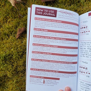 May include: A close-up of a journal with a pink and red cover. The journal is open to a page with instructions on how to use the journal. The page includes a section on tracking your menstrual cycle, mood, and physical and emotional experiences.