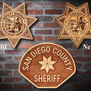 May include: Two wooden San Diego County Sheriff's badges, one old and one new. The old badge is a six-pointed star with the words "Deputy Sheriff" and "San Diego County Sheriff's Dept." The new badge is a simple shield shape with the words "San Diego County Sheriff".