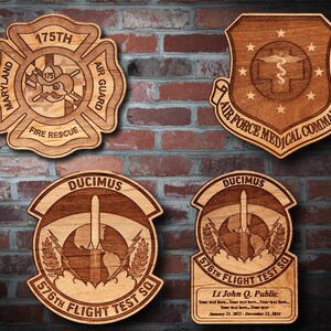 May include: Four wooden plaques with engraved text and designs. The top left plaque is a red, white, and blue Maryland Air Guard emblem with the text "Maryland 175th Air Guard Fire Rescue". The top right plaque is a brown Air Force Medical Command emblem with the text "Air Force Medical Command". The bottom left plaque is a brown plaque with the text "Ducimus 576th Flight Test Sq" and a rocket ship design. The bottom right plaque is a brown plaque with the text "Ducimus 576th Flight Test Sq" and a rocket ship design. The plaque also includes the text "Lt John Q. Public Your text here... January 21, 2022 - December 23, 2024".
