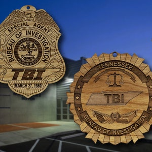 May include: Two wooden ornaments featuring the Tennessee Bureau of Investigation (TBI) logo. The first ornament is a shield-shaped badge with the text "Special Agent Bureau of Investigation" and "TBI" in the center. The second ornament is a circular design with the text "Tennessee Bureau of Investigation" and a map of Tennessee with "TBI" in the center.