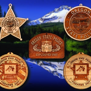 May include: Five wooden ornaments featuring the Oregon State Police logo. The ornaments are shaped like a star, a circle, and a rectangle. The star and circle ornaments feature the words "Oregon State Police" and the state seal. The rectangle ornament features the words "Oregon State Police" and "Explosives Unit". The other two ornaments feature the words "Oregon State Police" and "Firearms Unit".