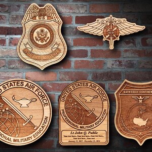 Könnte beinhalten: Fünf Holzplaketten mit gravierten Designs. Die oberste Plakette ist ein US-Spezialagenten-Abzeichen. Die zweite Plakette ist ein geflügeltes Spezialkriegsabzeichen. Die dritte und vierte Plakette sind Abzeichen der United States Air Force Professional Military Education. Die fünfte Plakette ist ein Barksdale Confinement-Abzeichen.
