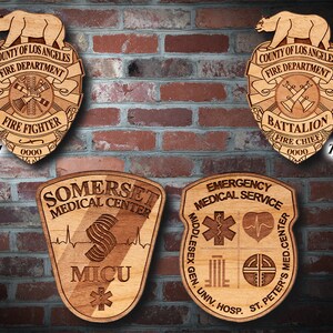 Könnte beinhalten: Vier Holzplaketten mit gravierten Designs. Die obere linke Plakette ist ein Feuerwehr-Abzeichen mit dem Text "COUNTY OF LOS ANGELES FIRE DEPARTMENT FIRE FIGHTER 0000". Die obere rechte Plakette ist ein Bataillonsführer-Abzeichen mit dem Text "COUNTY OF LOS ANGELES FIRE DEPARTMENT BATTALION FIRE CHIEF 0000". Die untere linke Plakette ist ein medizinisches Zentrum-Abzeichen mit dem Text "SOMERSET MEDICAL CENTER MICU". Die untere rechte Plakette ist ein Notfall-Rettungsdienst-Abzeichen mit dem Text "EMERGENCY MEDICAL SERVICE MIDDLESEX GEN. UNIV. HOSP. ST. PETER'S MED.CENTER".