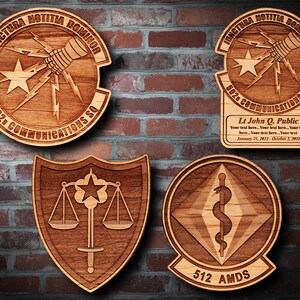 May include: Four wooden plaques with different military unit insignia. The top left plaque features a lightning bolt and a hand holding a microphone with the text "JUNCTURA NOTITIA DOMINOR 502D COMMUNICATIONS SQ". The top right plaque features the same insignia with the text "JUNCTURA NOTITIA DOMINOR 502 COMMUNICATIONS SQ" and a nameplate with the text "Lt John Q. Public Your text here... Your text here... Your text here... Your text here... Your text January 21, 2022 - October 3, 2023". The bottom left plaque features a shield with a scale of justice, a sword, and a star. The bottom right plaque features a diamond with a snake wrapped around a staff with the text "512 AMDS".