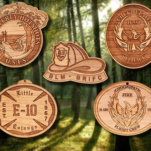 May include: Five wooden ornaments with different designs. The top left ornament is round and features a scene of a forest with the text "Monterey Hotshots U.S.F.S.". The top right ornament is round and features a phoenix with the text "Chuchupate H-530 UAS Helitack". The bottom left ornament is oval and features a cross with the text "Little E-10 1967 Tujunga". The bottom right ornament is oval and features a phoenix with the text "Chuchupate Fire H-530 Flight Crew USFS". The center ornament is shaped like a fire helmet and features the text "BLM BRIFC".