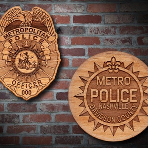 May include: Two wooden police badges. One is shield-shaped with "METROPOLITAN POLICE" and "OFFICER 000" engraved. The other is round with "METRO POLICE NASHVILLE DAVIDSON COUNTY" engraved. Both are set against a brick wall.