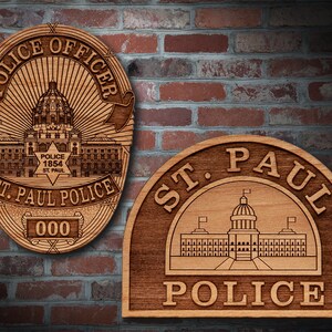 May include: Two wooden plaques with a St. Paul Police Department design. The first plaque is round and features a badge with a star, the words "Police 1854 St. Paul", and the text "St. Paul Police" in a banner. The second plaque is a half-circle with the words "St. Paul Police" and a building with a dome.