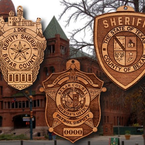 May include: Three wooden ornaments featuring the Bexar County Sheriff's Office logo. The first ornament is a traditional badge with the words "Deputy Sheriff Bexar County" and a star in the center. The second ornament is a star-shaped badge with the words "Bexar County Sheriff's Office Patrol Deputy" and a smaller badge in the center. The third ornament is a shield-shaped badge with the words "Sheriff The State of Texas County of Bexar".