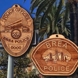 May include: Two wooden police badge ornaments. One is an oval shape with the words "POLICE OFFICER" and "BREA POLICE" and the number "0000". The other is a shield shape with "BREA POLICE" and a landscape design.