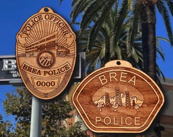 May include: Two wooden police badge ornaments. One is an oval shape with the words "POLICE OFFICER" and "BREA POLICE" and the number "0000". The other is a shield shape with "BREA POLICE" and a landscape design.