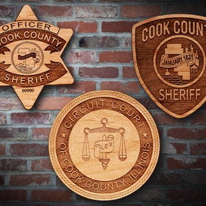 May include: Three wooden plaques with engraved designs. The top left plaque is a six-pointed star with the text "OFFICER COOK COUNTY SHERIFF 00000". The top right plaque is a shield with the text "COOK COUNTY SHERIFF JANUARY 1831". The bottom plaque is a circle with the text "CIRCUIT COURT OF COOK COUNTY ILLINOIS" and a scale of justice design.