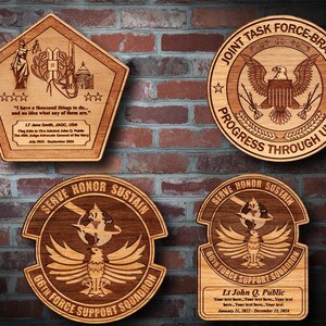 May include: Four wooden plaques with engraved designs. The top plaque is a pentagon shape with a quote and the text "LT Jane Smith, JAGC, USN." The bottom left plaque is round with the text "SERVE HONOR SUSTAIN 66TH FORCE SUPPORT SQUADRON." The bottom right plaque is round with the text "SERVE HONOR SUSTAIN 66TH FORCE SUPPORT SQUADRON Lt John Q. Public Your text here... Your text here... Your text here... Your text here... Your text here... January 21, 2022 - December 23, 2024." The top right plaque is round with the text "JOINT TASK FORCE-BRAVO PROGRESS THROUGH UNITY."