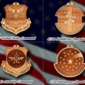 May include: Four wooden ornaments featuring the logos of different branches of the United States Air Force. The ornaments are in the shape of shields and circles with intricate designs and text. The logos include "AF Global Strike Command", "412th Healthcare Operations Sq", "Air Mobility Command", and "22D Intelligence Squadron".
