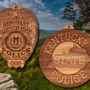 May include: Two wooden ornaments featuring the Kentucky State Police logo. The first ornament is a badge with the words "Sergeant Kentucky State Police" and "000". The second ornament is a round shape with the words "Kentucky State Police" and an outline of the state of Kentucky.