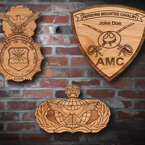 Könnte beinhalten: Drei Holzplaketten mit gravierten Designs. Die obere Plakette ist schildförmig mit dem Text "PARSONS MOUNTED CAVALRY" und "John Doe" und "AMC". Die mittlere Plakette ist kreisförmig mit dem Text "SECURITY POLICE DEPARTMENT OF THE AIR FORCE" und "UNITED STATES OF AMERICA" und "A1234". Die untere Plakette ist ein geflügeltes Globus-Design mit einem Stern.