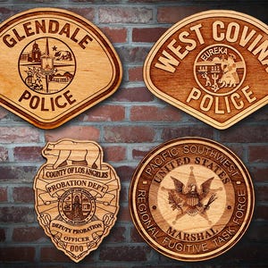 Peut inclure: Quatre plaques en bois avec différents logos d'agences d'application de la loi. La plaque en haut à gauche est pour le service de police de Glendale, la plaque en haut à droite est pour le service de police de West Covina, la plaque en bas à gauche est pour le service de probation du comté de Los Angeles, et la plaque en bas à droite est pour la force spéciale de recherche de fugitifs de la région du Pacifique Sud-Ouest.