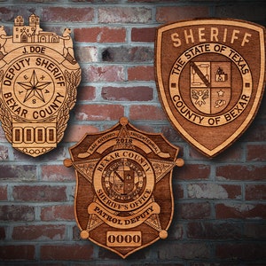 May include: Three wooden sheriff badges with the text "Sheriff", "Deputy Sheriff", and "Sheriff's Office Patrol Deputy" on them. The badges are all brown and have a star shape. The badges are on a brick wall.