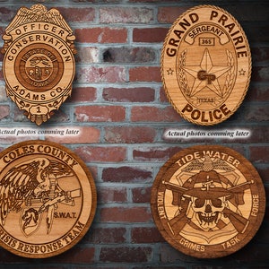 May include: Four wooden plaques featuring different law enforcement designs. The top left plaque is a badge with the text "Officer Conservation Adams Co. 1". The top right plaque is oval shaped with the text "Grand Prairie Police 365". The bottom left plaque is round with the text "Coles County Crisis Response Team S.W.A.T.". The bottom right plaque is round with the text "Tide Water Violent Crimes Task Force".