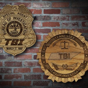 May include: Two wooden plaques with the Tennessee Bureau of Investigation (TBI) logo. The top plaque is a shield shape with the words "Special Agent Bureau of Investigation" and "TBI" with the date "March 27, 1980". The bottom plaque is a circular shape with the words "Tennessee Bureau of Investigation" and "TBI" with a scale of justice and the words "Truth, Bravery, Integrity".
