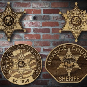 May include: Four wooden plaques with the Orange County Sheriff's Department logo. The top two plaques are shaped like a six-pointed star and read "DEPUTY SHERIFF" and "SERGEANT DEPUTY SHERIFF". The bottom left plaque is round and reads "ORANGE COUNTY SHERIFF'S DEPARTMENT EST. 1889". The bottom right plaque is shaped like a hexagon and reads "ORANGE COUNTY SHERIFF".