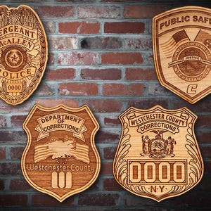 May include: Four wooden plaques with engraved designs. The top left plaque is a police badge with the text "SERGEANT McALLEN POLICE 0000". The top right plaque is a public safety badge with the text "PUBLIC SAFETY UNIVERSITY OF TENNESSEE JUSTICE AT CHATTANOOGA C". The bottom left plaque is a department of corrections badge with the text "DEPARTMENT OF CORRECTIONS Westchester County". The bottom right plaque is a Westchester County Corrections badge with the text "WESTCHESTER COUNTY CORRECTIONS 0000 NY".