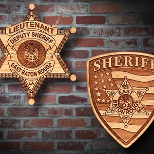 May include: Two wooden sheriff's badges. The first is a five-pointed star with the text "Lieutenant Deputy Sheriff State of Louisiana East Baton Rouge LA". The second is a shield shape with the text "Sheriff" and a smaller star with the text "East Baton Rouge".