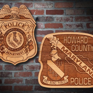 May include: Two wooden police badges, one shield-shaped and the other a modified badge shape, are displayed against a brick wall. The shield badge features an eagle, the words "POLICE," "HOWARD COUNTY," and "MARYLAND DEPARTMENT." The other badge reads "HOWARD COUNTY MARYLAND POLICE."