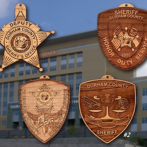 May include: Four wooden ornaments featuring the Durham County Sheriff's Office logo. The ornaments are shaped like a star, a shield, and two different versions of a badge. The star ornament says "Deputy Durham County Sheriff's Office". The shield ornament says "Sheriff Durham County Honor Duty Service 1881". The badge ornaments say "Durham County Sheriff's Office" and "Durham County Sheriff #2 Criminal Investigations Division".
