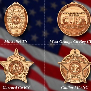 May include: Four wooden ornaments featuring law enforcement badges. The top left ornament is a brown oval with the text "Police City of Mt. Juliet TN". The top right ornament is a brown circle with the text "West Orange County Regional CERT Community Emergency Response Team". The bottom left ornament is a brown five-pointed star with the text "Deputy Garrard County Kentucky Sheriff's Association". The bottom right ornament is a brown five-pointed star with the text "Sheriff's Office Guilford County Sheriff".