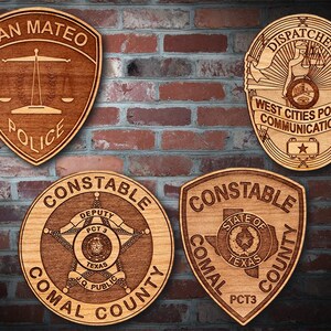 May include: Four wooden plaques with engraved police badges. The top left plaque reads "San Mateo Police". The top right plaque reads "Dispatcher West Cities Police Communications". The bottom left plaque reads "Constable Comal County Deputy PCT 3 J.Q. Public". The bottom right plaque reads "Constable Comal County State of Texas PCT 3".