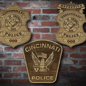 May include: Three wooden Cincinnati Police badges are displayed against a brick wall. Two badges are shield-shaped, one labeled "Detective." The third badge is a dark brown rectangle with an eagle and American flag.