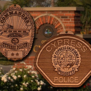 May include: Two wooden ornaments featuring the Cypress, California Police Department logo. The first ornament is a traditional police badge with the words "Commander" and "Cypress Police" inscribed. The second ornament is a shield-shaped badge with the words "Cypress, City of Cypress, California, 1956, Progress, Police" inscribed.