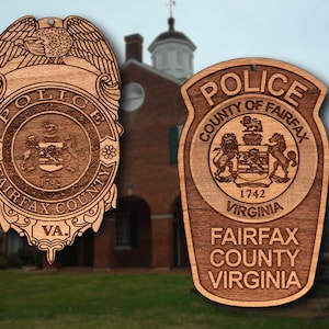 May include: Two wooden ornaments shaped like police badges. The first badge features a stylized eagle with the text "POLICE FAIRFAX COUNTY VA." The second badge features a coat of arms with the text "POLICE COUNTY OF FAIRFAX 1742 VIRGINIA FAIRFAX COUNTY VIRGINIA."