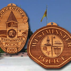 May include: Two wooden ornaments featuring the Westminster Police Department logo. The first ornament is shaped like a badge and features the words "Police Officer" and "Westminster Police 000". The second ornament is a round, wooden plaque with the words "Westminster Police" in a circle.