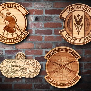 May include: Four wooden plaques with engraved military insignia. The top left plaque reads "CONTEGO ☆ 5TH SECURITY FORCES SQ". The top right plaque reads "COMMANDO ☆ 6TH SPECIAL OPS SQ". The bottom left plaque features a stylized eagle with wings spread. The bottom right plaque reads "TO HONOR WITH DIGNITY USAF HONOR GUARD".