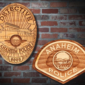 May include: Two wooden Anaheim Police badges. One is oval-shaped, with "DETECTIVE" at the top and "ANAHEIM POLICE" at the bottom. The other is a semi-circular badge with "ANAHEIM POLICE" at the bottom. Both are light brown.