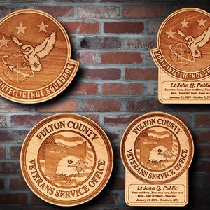 May include: Four wooden plaques with engraved designs. The top two plaques feature a stylized owl with a globe and the text "204th Intelligence Squadron". The bottom two plaques feature a stylized eagle with the text "Fulton County Veterans Service Office". Each plaque includes a personalized name and dates.