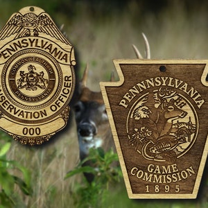May include: Two wooden Pennsylvania state-themed ornaments. One is a badge-shaped ornament with "PENNSYLVANIA CONSERVATION OFFICER" and "000" engraved. The other is a keystone-shaped ornament with "PENNSYLVANIA GAME COMMISSION 1895" and a wildlife scene.