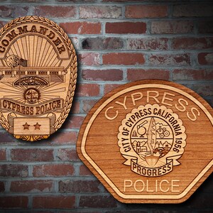 May include: Two wooden Cypress Police badges are displayed against a brick wall. The first badge is oval-shaped, with the word "Commander" at the top and "Cypress Police" at the bottom. The second badge is shield-shaped, with "Cypress California 1956" and "Police" on it.
