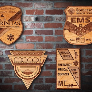 Könnte beinhalten: Vier Holzplaketten mit unterschiedlichen Designs. Die obere linke Plakette ist braun und trägt den Text "EMERGENCY MEDICAL SERVICES TRINITAS Regional Medical Center" mit einem Stern-des-Lebens-Symbol. Die obere rechte Plakette ist braun und trägt den Text "Somerset MEDICAL CENTER EMS EST. 1982" mit einem Stern-des-Lebens-Symbol und einer amerikanischen Flagge. Die untere linke Plakette ist braun und trägt den Text "New Jersey's MONOC INTEGRITY PROFESSIONALISM EXCELLENCE" mit einer Sonnenuntergangsgrafik. Die untere rechte Plakette ist braun und trägt den Text "MEDICAL CENTER EMERGENCY MEDICAL SERVICES JERSEY CITY N.J. MC_JE_" mit einem Stern-des-Lebens-Symbol und einer Grafik der Freiheitsstatue.