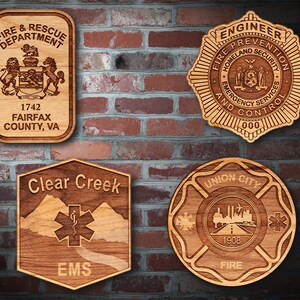May include: Five wooden plaques with engraved designs. The top left plaque is a brown rectangle with the text "FIRE & RESCUE DEPARTMENT" and "1742 FAIRFAX COUNTY, VA". The top right plaque is a brown star with the text "ENGINEER FIRE PREVENTION HOMELAND SECURITY EMERGENCY SERVICES AND CONTROL 000". The bottom left plaque is a brown hexagon with the text "Clear Creek" and "EMS". The bottom right plaque is a brown circle with the text "UNION CITY 1908 FIRE".