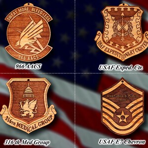 May include: Four wooden ornaments featuring US Air Force insignia. The top left ornament is a brown shield with a white eagle and the text "SWEAT MORE, BLEED LESS" and "966 AACS". The top right ornament is a brown shield with a white wreath and crossed arrows and the text "USAF EXPEDITIONARY CENTER". The bottom left ornament is a brown shield with a white eagle and the text "316TH MEDICAL GROUP". The bottom right ornament is a brown shield with a white star and the text "MSGT JOHN Q. PUBLIC 0000".