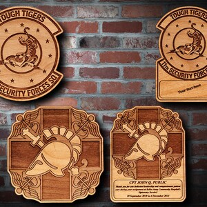 May include: Four wooden plaques with laser-engraved designs. The top left plaque features a tiger and the text "TOUGH TIGERS 11th SECURITY FORCES SQ". The top right plaque features a tiger and the text "TOUGH TIGERS 11th SECURITY FORCES SQ" with a blank space for text below. The bottom left plaque features a stylized helmet and sword design. The bottom right plaque features a stylized helmet and sword design with the text "CPT JOHN Q. PUBLIC Thank you for your dedicated leadership and compassionate patient care during your assignment at Keller Army Community Hospital's Optometry Service. 19 September 2019 to 4 December 2023".