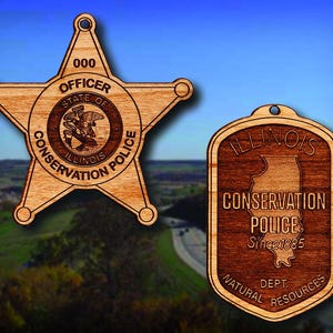 May include: Two wooden ornaments featuring the Illinois Conservation Police logo. The first is a five-pointed star with the text "000 OFFICER STATE OF ILLINOIS CONSERVATION POLICE". The second is a shield shape with the text "ILLINOIS CONSERVATION POLICE Since 1885 DEPT. NATURAL RESOURCES".