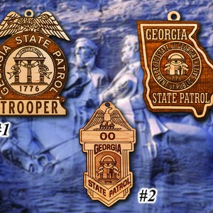 May include: Three wooden ornaments shaped like the state of Georgia, a badge, and a shield. The ornaments are decorated with the Georgia State Patrol logo and the words "Georgia State Patrol".