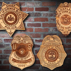 May include: Four wooden police badges with different designs and text. The badges are displayed on a brick wall. The text on the badges includes "Chief California Boro Police", "Chaplain New York State Chaplain Task Force NYS", "US National Nuclear Security Administration Federal Agent 0000", and "University of Tennessee Police Officer Chattanooga".