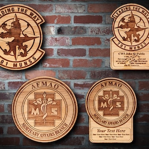 Könnte beinhalten: Vier Holzplaketten mit gravierten Designs. Zwei Plaketten zeigen einen Löwen und einen Kompass mit dem Text "LEADING THE WAY 701 MUNSS". Eine Plakette zeigt ein Kreuz mit einer Schlange und dem Text "AFMAO MORTUARY AFFAIRS BRANCH". Die letzte Plakette zeigt das gleiche Kreuz und die gleiche Schlange mit dem Text "AFMAO MORTUARY AFFAIRS BRANCH" und einem Platz für personalisierten Text.